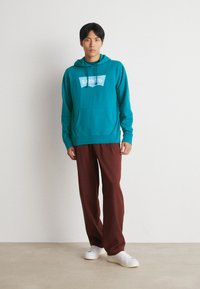Turquoise hoodie met witte Levi's-logo, kangoeroezak, gecombineerd met bruine broek en witte sneakers op een lichte achtergrond.