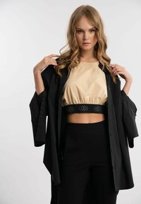 Sort oversized skjorte med knapdetaljer, båret over en beige cropped top med strammet elastisk kant og sorte accenter. Model poserer.
