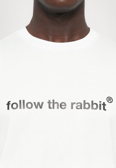 Valge puuvillane T-särk, millel on julge, tumehall tekst "follow the rabbit®", mis on kirjutatud rinna peale. Lihtne, klassikaline disain.