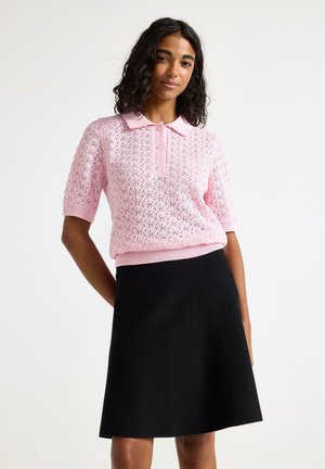 Kiharatukkainen nainen, joka pukeutuneena vaaleanpunaiseen neulottuun poolopaitaan ja mustaan polvenpituiseseen hameeseen, poseeraa yksinkertaista valkoista taustaa vasten.