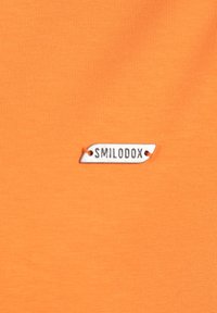 Oranje stof met een gladde textuur, voorzien van een wit rechthoekig label met de tekst "SMILODOX" in zwarte print, bevestigd met een klein oranje stiksel.