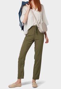Pantaloni verde khaki con vita elastica, abbinati a una blusa bianca larga e a un leggero giubbotto di jeans blu drappeggiato sulla spalla. Espadrillas beige completano il look, insieme a una piccola borsa a tracolla rotonda color crema.