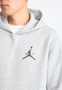 Felpa grigia con cappuccio realizzata in tessuto morbido, caratterizzata da un logo Jumpman nero con il testo "A" e "R", che mostra un design semplice e sportivo.