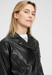 Schwarze Leder-Bikerjacke mit Reißverschluss-Taschen und Druckknopfkragen. Besitzt eine taillierte Silhouette und eine glatte Textur. Dicke Nähte sichtbar.