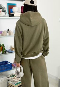 Olívovo zelená oversized mikina s kapucňou, rebrovaným lemom a spadnutými plecami, v kombinácii s koženkovými nohavicami. Drží biele slúchadlá.