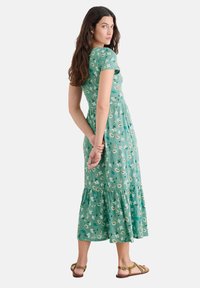 Rochie florală verde cu mâneci scurte, tiv în straturi, fabricată dintr-un material moale. Model floral alb cu frunze pe întreaga suprafață. Pur tată cu sandale maro.