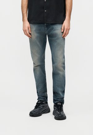 Homme portant un jean bleu délavé, une chemise noire boutonnée et des baskets noires Diesel, debout devant un fond blanc.