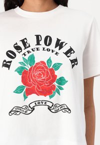 Fehér póló, amelyen egy nagy piros rózsa grafika látható, zöld levelekkel, fekete szöveggel a "ROSE POWER TRUE LOVE" felirattal felül és a "SPIRIT OF LOVE THE SOUL" felirattal alul.