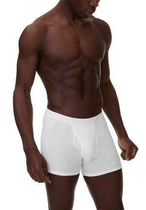 Uomo muscoloso che indossa boxer bianchi, in piedi davanti a uno sfondo bianco uniforme, con lo sguardo rivolto verso il basso e i pugni serrati.