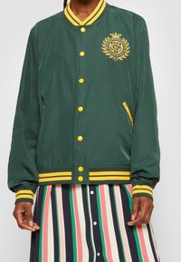 Giacca bomber verde con accenti gialli, bottoni a pressione, polsini a coste e un emblema logo. Abbinata a una gonna a righe sotto.
