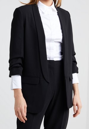 Femme portant un blazer noir cintré et un pantalon assorti sur une chemise blanche à boutons, debout devant un fond clair uni.