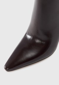 Bottine en cuir brun foncé à bout pointu avec une finition lisse et brillante sur un fond clair.