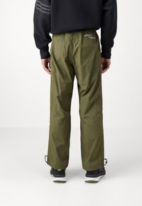 Bakifrån av en person som bär olivgröna Adidas Originals-byxor, svart sweatshirt och svarta sneakers med vita sulor mot en vit bakgrund.