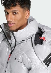 Ασημί μπουφάν puffer με ψηλό γιακά, τσέπες με φερμουάρ και ελαστικές λεπτομέρειες. Διαθέτει μαύρα και κόκκινα μεταλλικά στοιχεία με λογότυπο στο στήθος.
