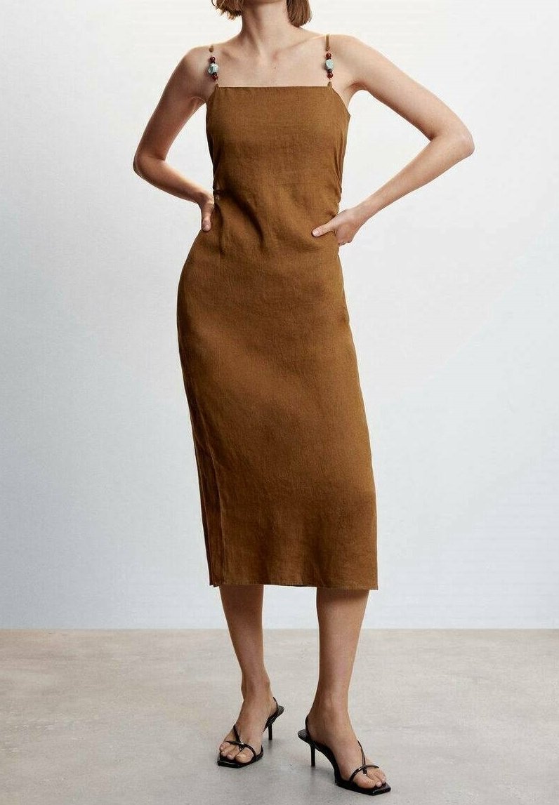 Femme portant une robe midi marron sans manches avec des bretelles décoratives en perles et des sandales noires à talons hauts, debout les mains sur les hanches.