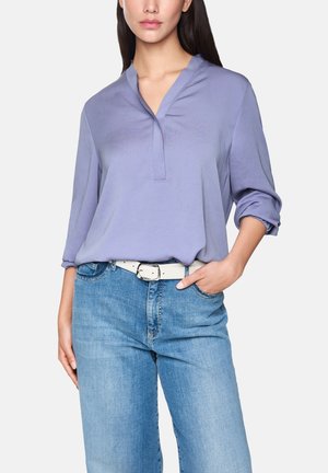 Woman wearing a light purple V-neck blouse with rolled-up sleeves tucked into light blue jeans with a white belt, hand in pocket.