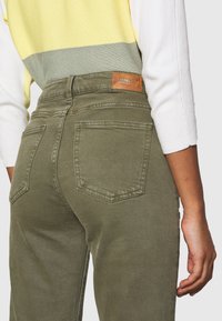 Pantalon taille haute vert olive à texture lisse, doté de deux poches arrière et d'une étiquette en cuir de marque à la ceinture.