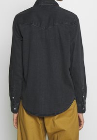 Camicia in denim nera con maniche lunghe, colletto a punta e dettaglio sulla parte posteriore. Presenta polsini a bottone e orlo curvo. Indossata con pantaloni beige.