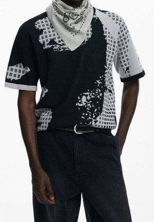 Mand iført en kortærmet sweater med sort og gråt abstrakt mønster, sorte bukser med bælte og et lyst gråt mønstret halstørklæde.