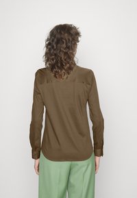 Camicetta marrone a maniche lunghe con una texture liscia, dettaglio di spalla, e polsini con bottoni, indossata con pantaloni verde chiaro.