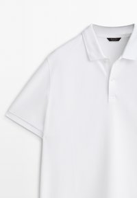 Polo blanc avec un col classique, manches courtes et tissu texturé. Patte de boutonnage à trois boutons près de l'encolure.