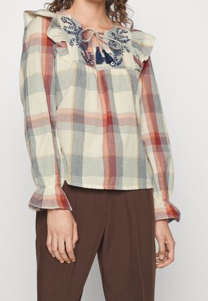 Kobieta w kraciastej bluzce z długim rękawem, z haftowanym dekoltem, bufkami na ramionach i troczkami z chwostami, w połączeniu z brązowymi spodniami.