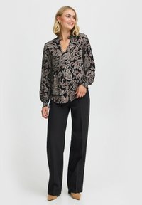 Blusa nera con motivo paisley rosa e crema, polsini a sbuffo e scollatura a V, abbinata a pantaloni neri larghi.