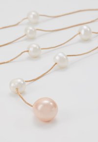 Collar de tono dorado que presenta una perla irregular blanca y una de color melocotón, con un diseño de cadena retorcida, mostrando texturas suaves y brillantes.