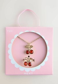 Guld-tone kirsebær halskæde og øreringe med røde ædelstene detaljer, pakket i en lyserød æske med et rundt udsnit og bølgende kant.