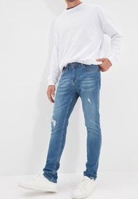 Jean en denim bleu clair délavé avec une coupe slim, présentant des zones décolorées et des détails déchirés, associé à une chemise blanche à manches longues.