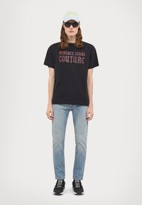 Svart t-skjorte med "VERSACE JEANS COUTURE" grafikk, lyseblå jeans, svarte joggesko med hvit tekst, grå caps og solbriller.