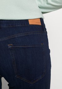 Jeans de mezclilla azul oscuro con parche de cuero marrón claro, costuras de contraste y diseño tradicional de bolsillo trasero. Detalle en primer plano.