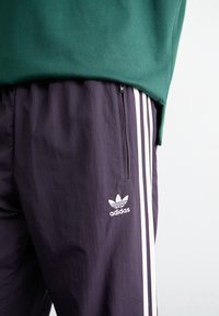 Lila träningsbyxor med vita sidostripes, dragkedjeficka och Adidas-logotyp på vänster ben. Bärs med mörkgrön sweatshirt ovanför.