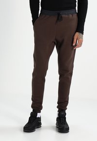 Pantalones de chándal marrones con una cintura elástica negra, diseño de pierna ajustada y cordón. Combinados con zapatillas deportivas negras y calcetines blancos.