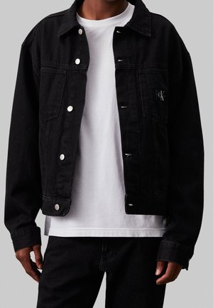 Mand iført en sort denimjakke med hvide knapper over en ensfarvet hvid t-shirt og sorte bukser mod en grå baggrund.
