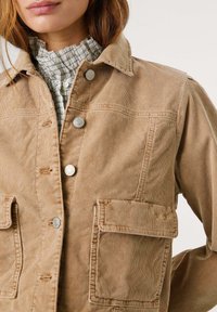 Tan corduroyjas met een knoopsluiting aan de voorkant, twee grote borstzakken en een kraag. Gedragen over een geruite, met kant afgewerkte kraagshirt.