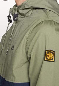 Grön huvad jacka med dragsko, svart och gröna detaljer samt en tydlig gul broderad logga på ärmen.