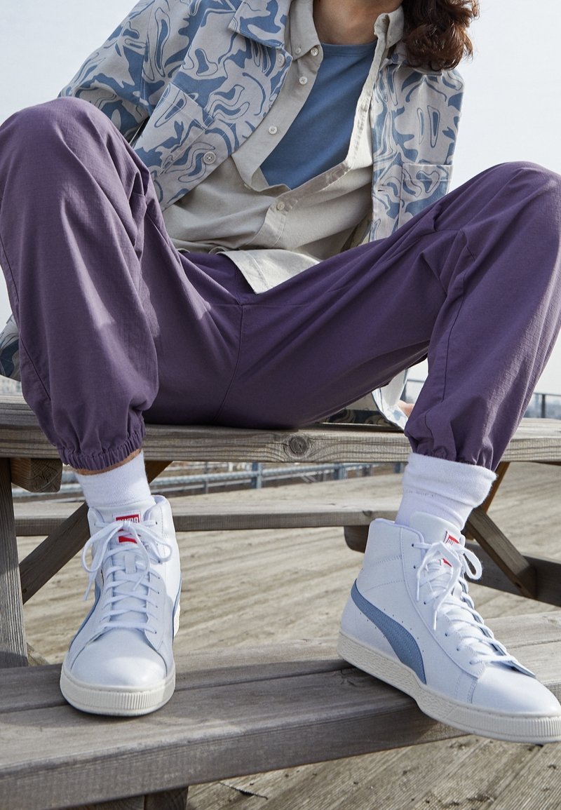 Bílé vysoké tenisky s světle modrými akcenty, nošené s fialovými zúženými kalhotami a vzorovanou světle modrou a bílou bundou.