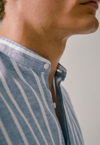 Lähikuva henkilön kaulasta ja leuasta, joka käyttää sinivalkoraidallista napitettua paitaa, jossa on pystykrae.