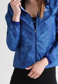 Veste de sport bleue avec un motif en vagues, dotée d'une fermeture éclair à l'avant, de poignets côtelés et d'une capuche. Fabriquée en matériau léger et respirant.