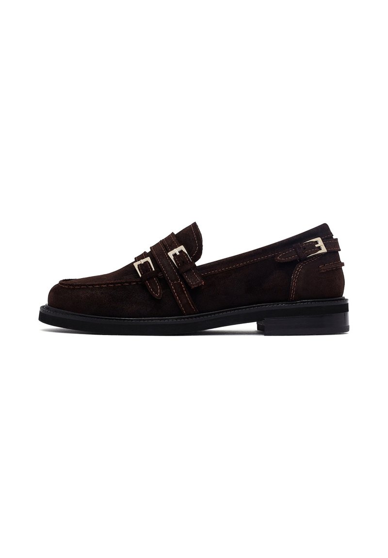 Mocassino in suede marrone con doppie cinghie e dettagli in fibbia argentata. Suola in gomma nera. Punta arrotondata e cuciture a contrasto.