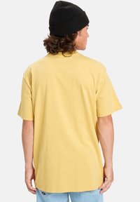 Žluté bavlněné tričko s volným střihem, krátkými rukávy a kulatým výstřihem. Nosené s světle modrými džíny a černou čepicí.