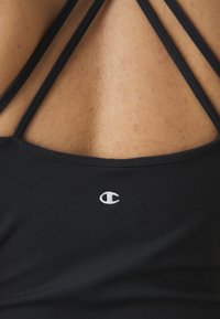 Crni sportski top bez rukava s tankim križnim trakama na leđima i malim bijelim logotipom na donjoj lijevoj strani. Glatka, rastezljiva tkanina.