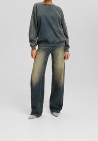 Sudadera gris de gran tamaño con puños acanalados, combinada con vaqueros de pierna ancha en azul desgastado. Unos zapatos azul claro completan el atuendo.