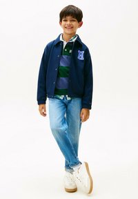Ragazzo sorridente in piedi con le gambe incrociate, che indossa una giacca navy con lettera "H", una maglietta da rugby a righe, jeans azzurri chiari e sneaker bianche su sfondo bianco.