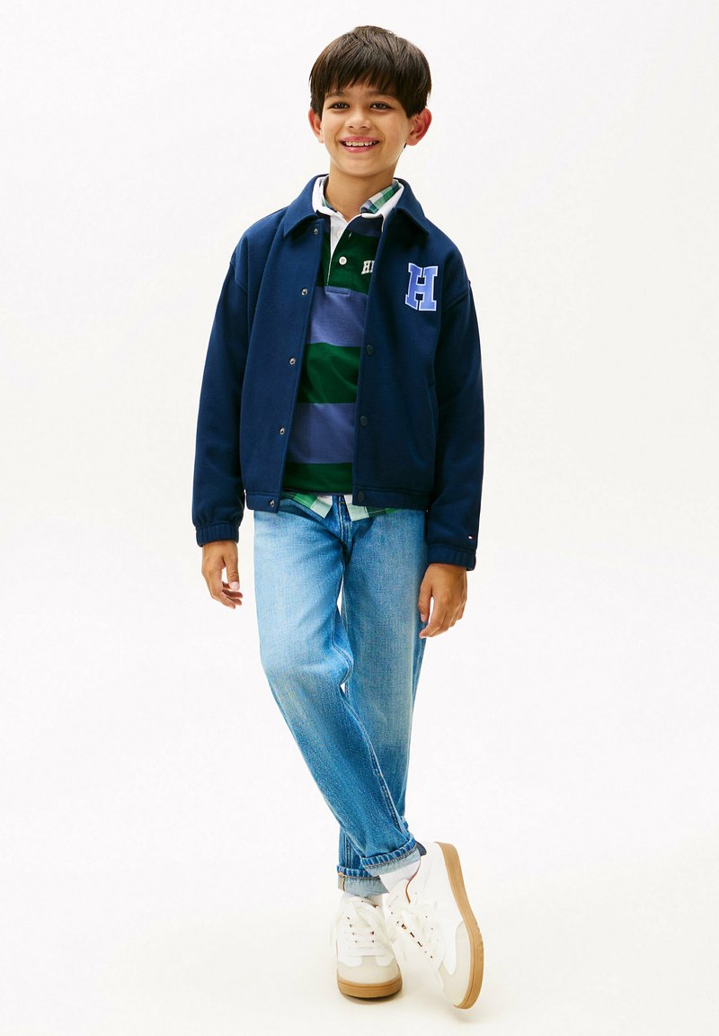 Ragazzo sorridente in piedi con le gambe incrociate, che indossa una giacca navy con lettera "H", una maglietta da rugby a righe, jeans azzurri chiari e sneaker bianche su sfondo bianco.