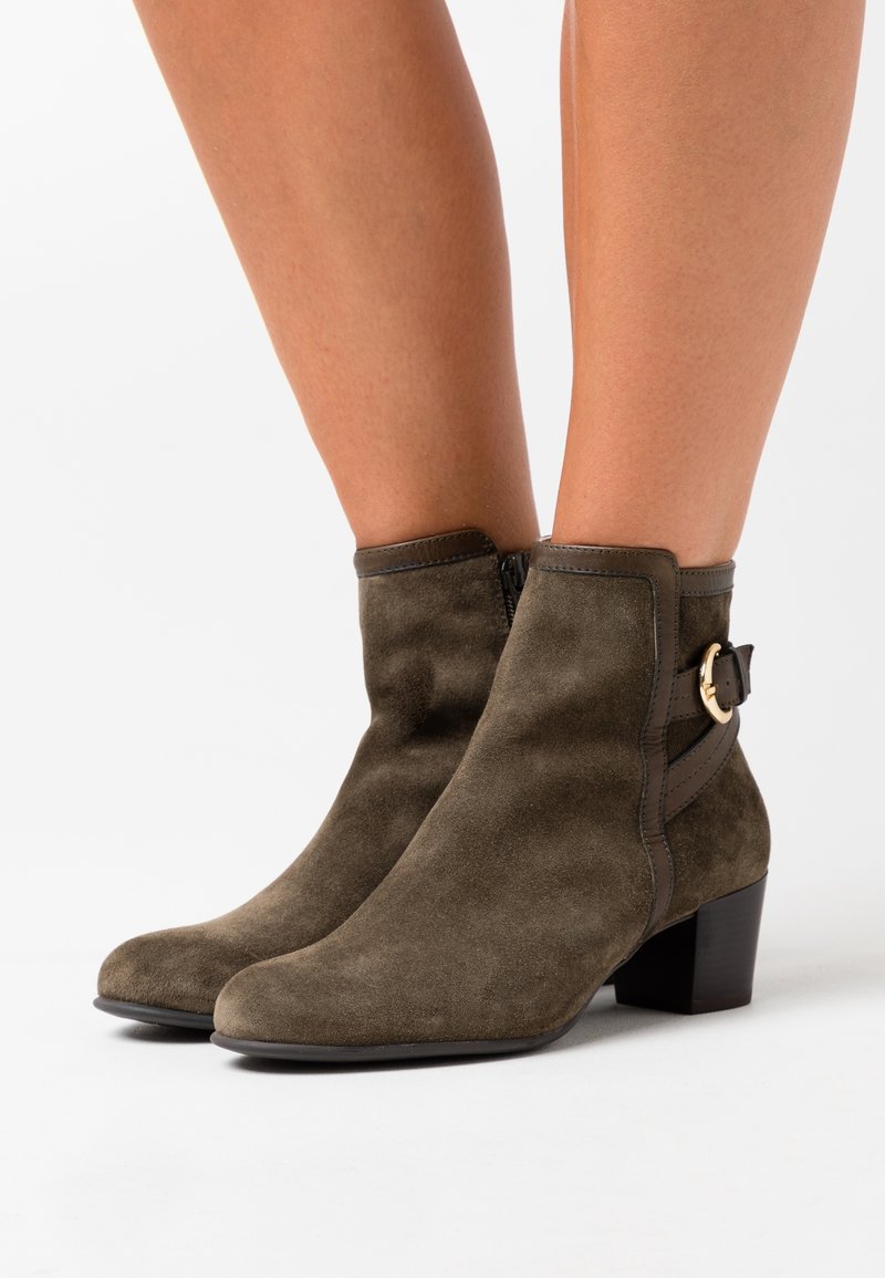 Bottes en daim olive montant à la cheville avec un talon bloc bas, une fermeture éclair latérale et une sangle décorative avec boucle dorée portées sur des jambes nues.