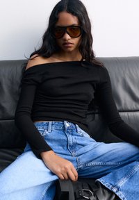 Zwarte off-shoulder lange mouw top gecombineerd met lichtblauwe hoge taille jeans en chunky zwarte plateau schoenen; zittend op een zwarte leren bank.