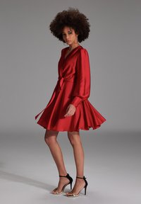 Robe en satin rouge avec des manches longues et une taille ceinturée, dotée d'une jupe évasée. Des sandales à talons hauts noires et dorées complètent la tenue.