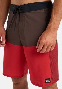 Homme portant un short de bain marron et rouge avec une taille noire et un cordon de serrage, se tenant avec les mains détendues sur les côtés.